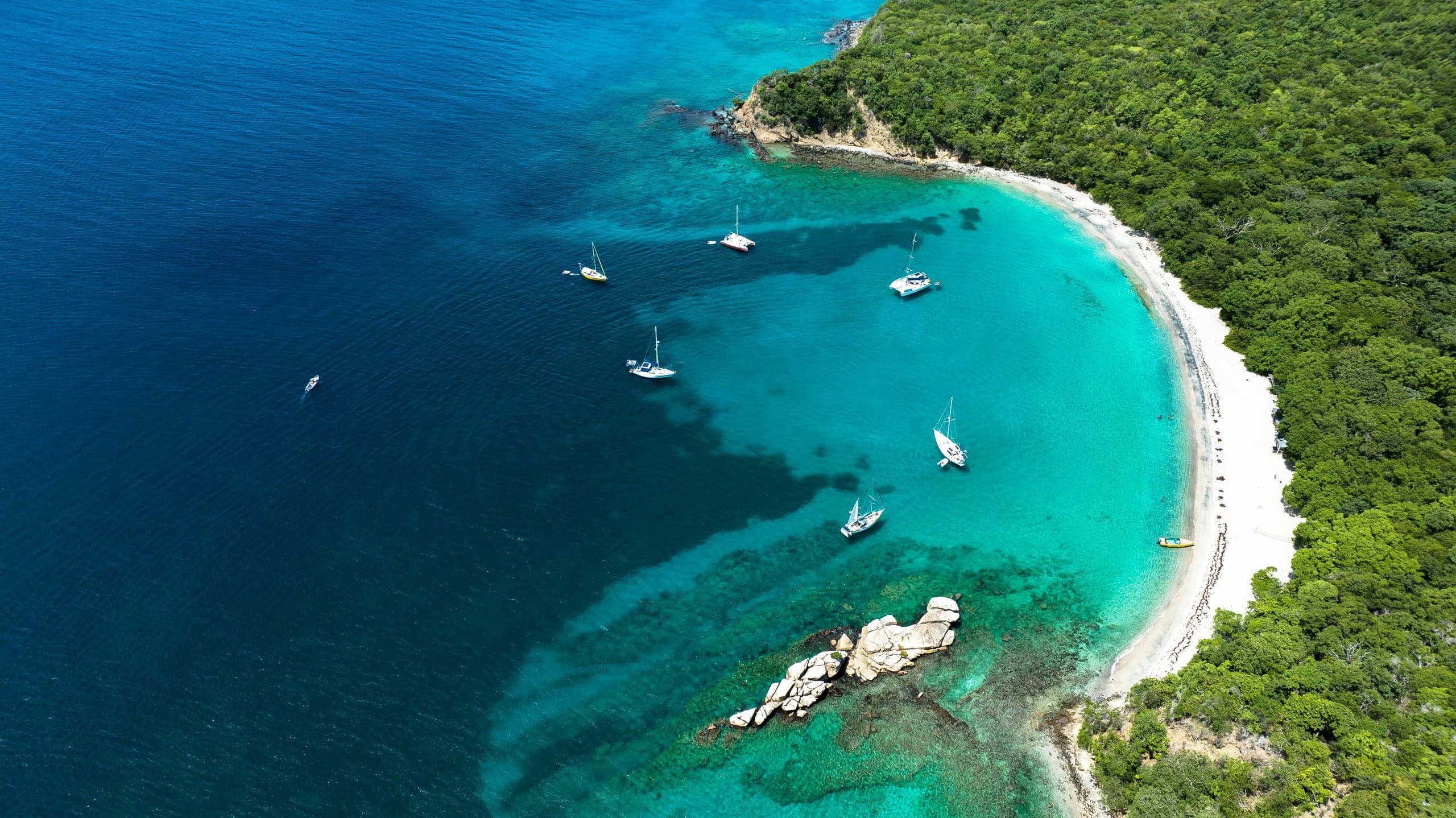 A wooded cove with sailboats at anchor in turquoise water, seen from the air