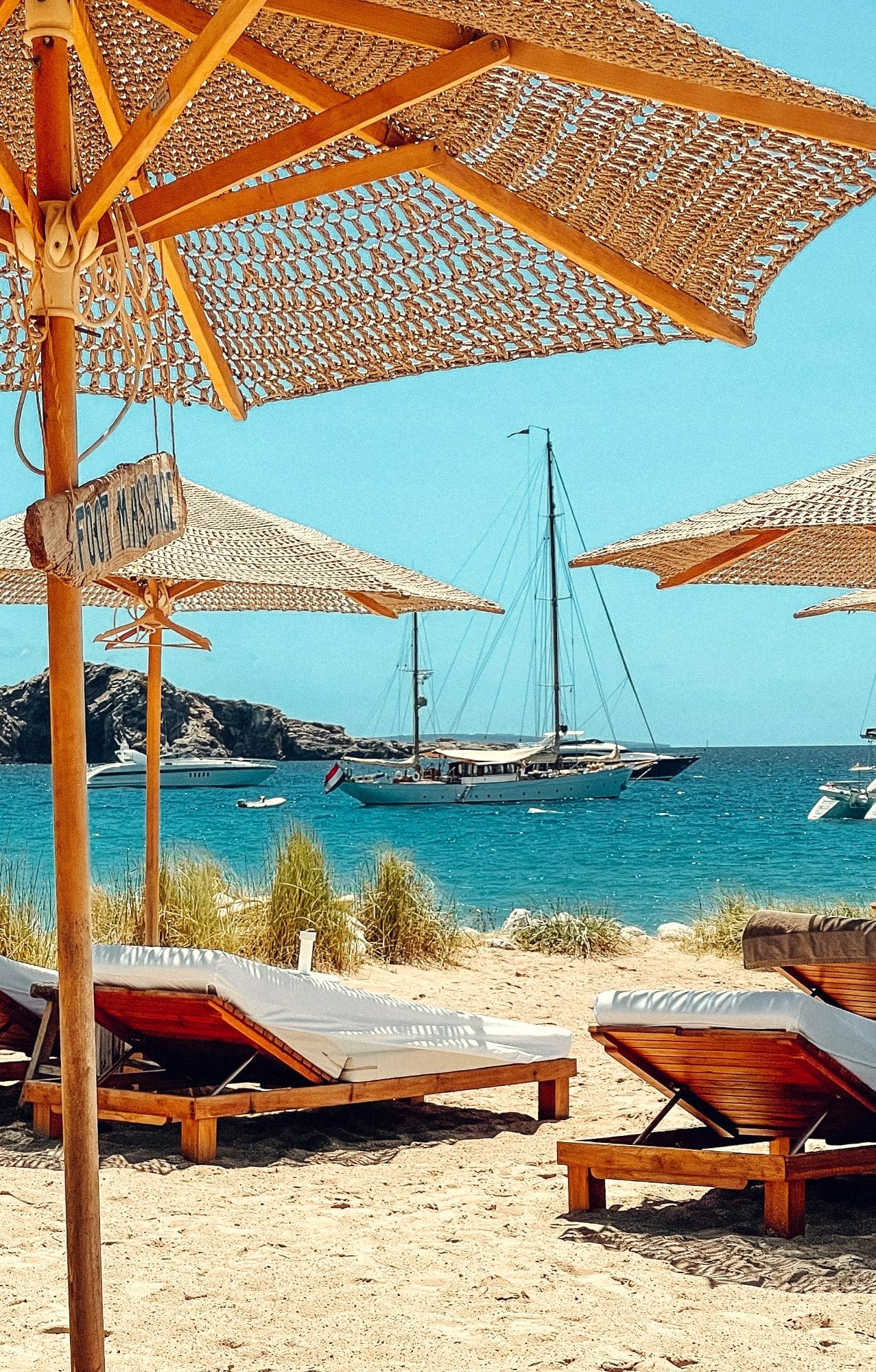 Straw umbrellas and loungers on a beach, yachts at anchor beyond